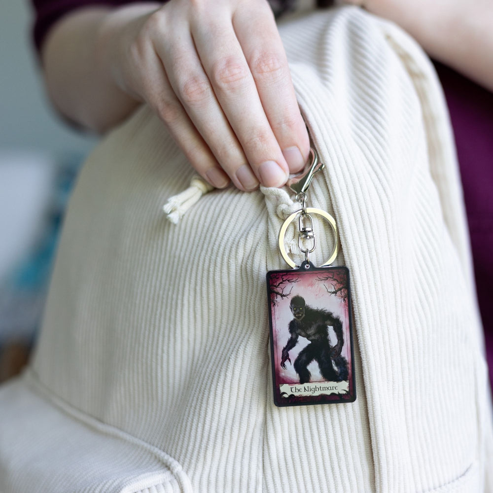 Acrylic keychain of a red tarot card that says ‘The Nightmare’ across the bottom  clipped to a white backpack. The card has creepy branches at the top and a monster in frame. 
