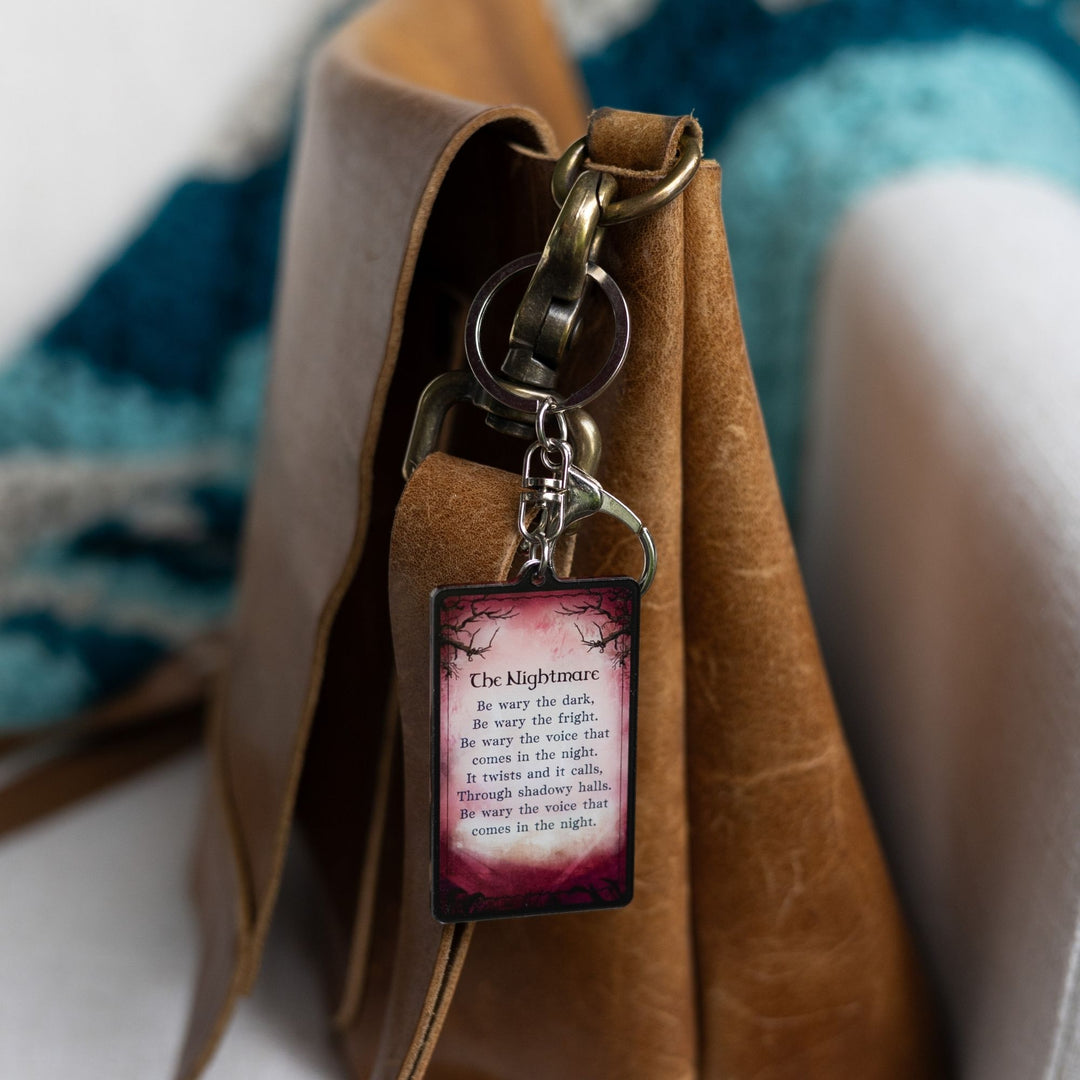 Acrylic keychain of a red tarot card with a poem that starts ‘The Nightmare. Be wary the dark, be wary the fright. Be wary the voice that comes in the night…’ clipped to a leather purse.’
