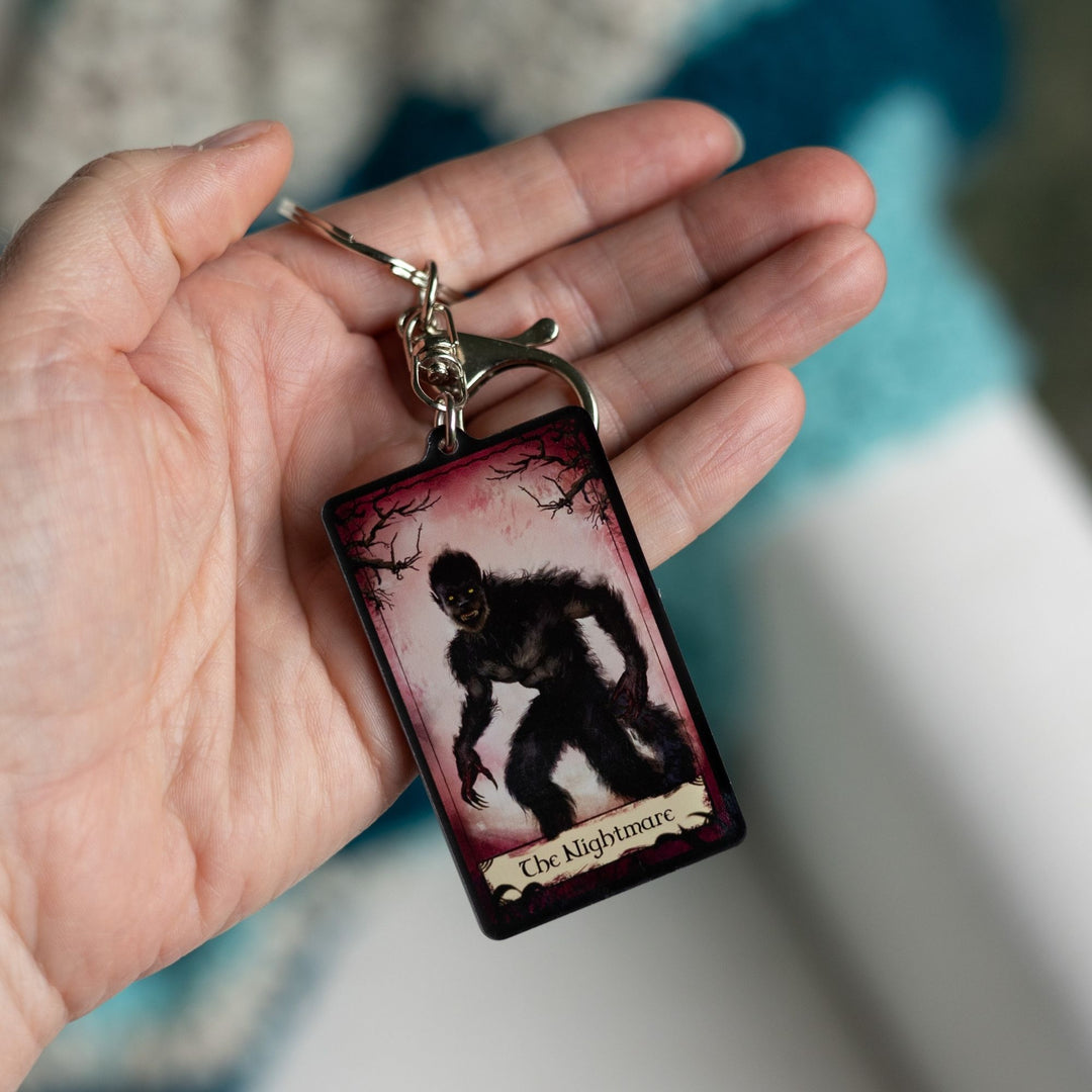 Acrylic keychain of a tarot card that says ‘The Nightmare’ across the bottom. The card has a red background with creepy branches at the top and the Nightmare in frame. Held in a palm.

