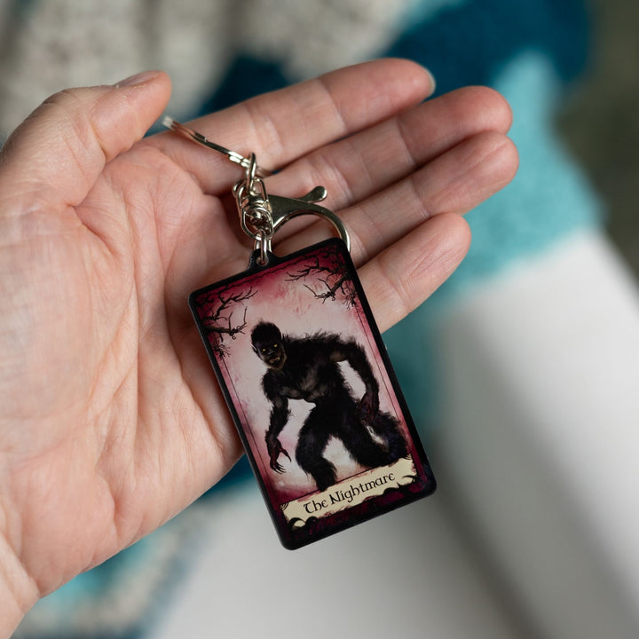 Acrylic keychain of a tarot card that says ‘The Nightmare’ across the bottom. The card has a red background with creepy branches at the top and the Nightmare in frame. Held in a palm.
