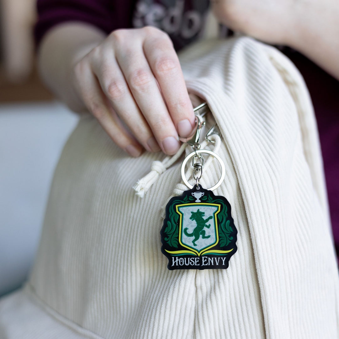  Keychain of a crest with a two headed wolf with green leaves on the side, a trophy on top, and House of Envy in white letters at the bottom clipped to a white backpack.
