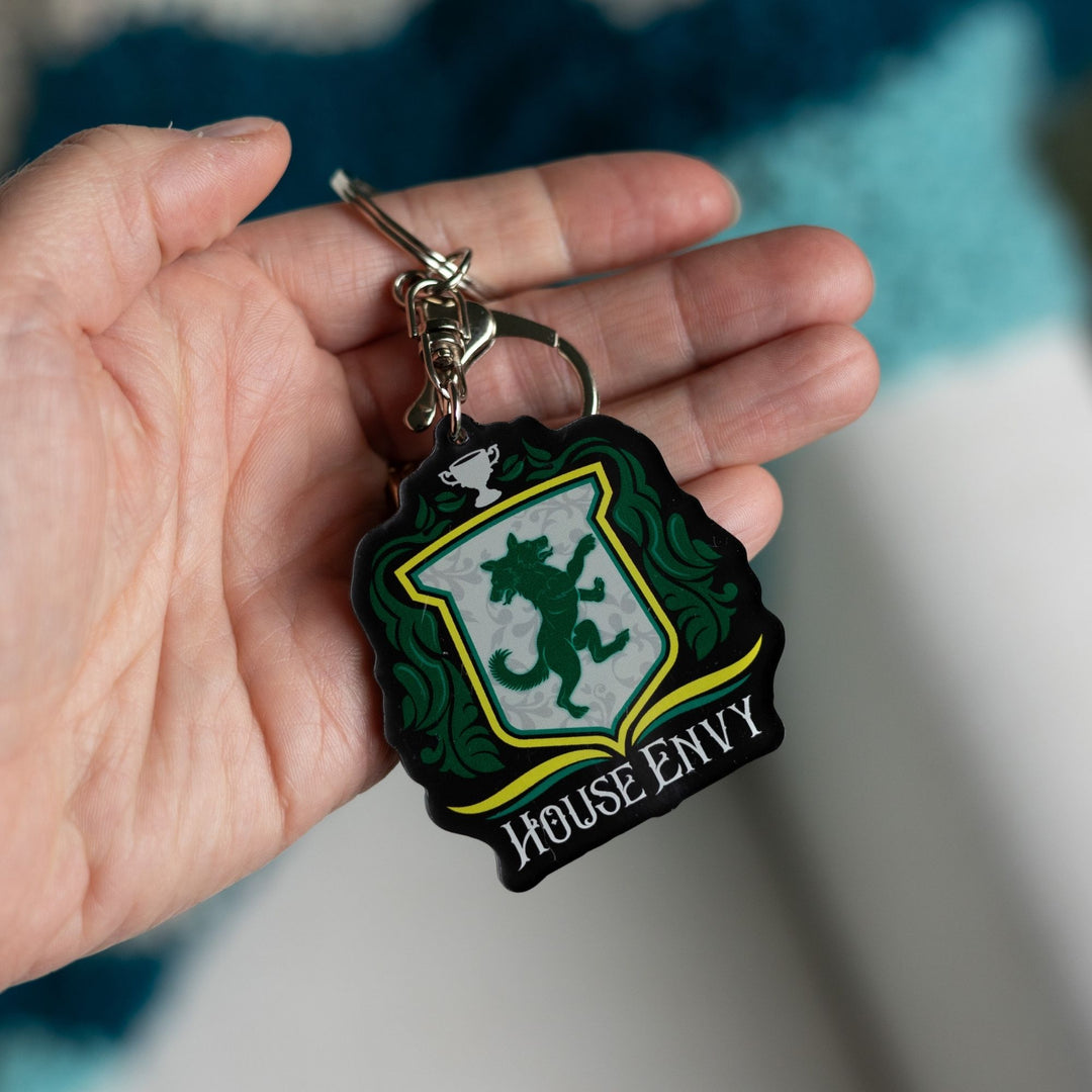 Keychain of a crest with a two headed wolf with green leaves on the side, a trophy on top, and House of Envy in white letters at the bottom, held in a palm