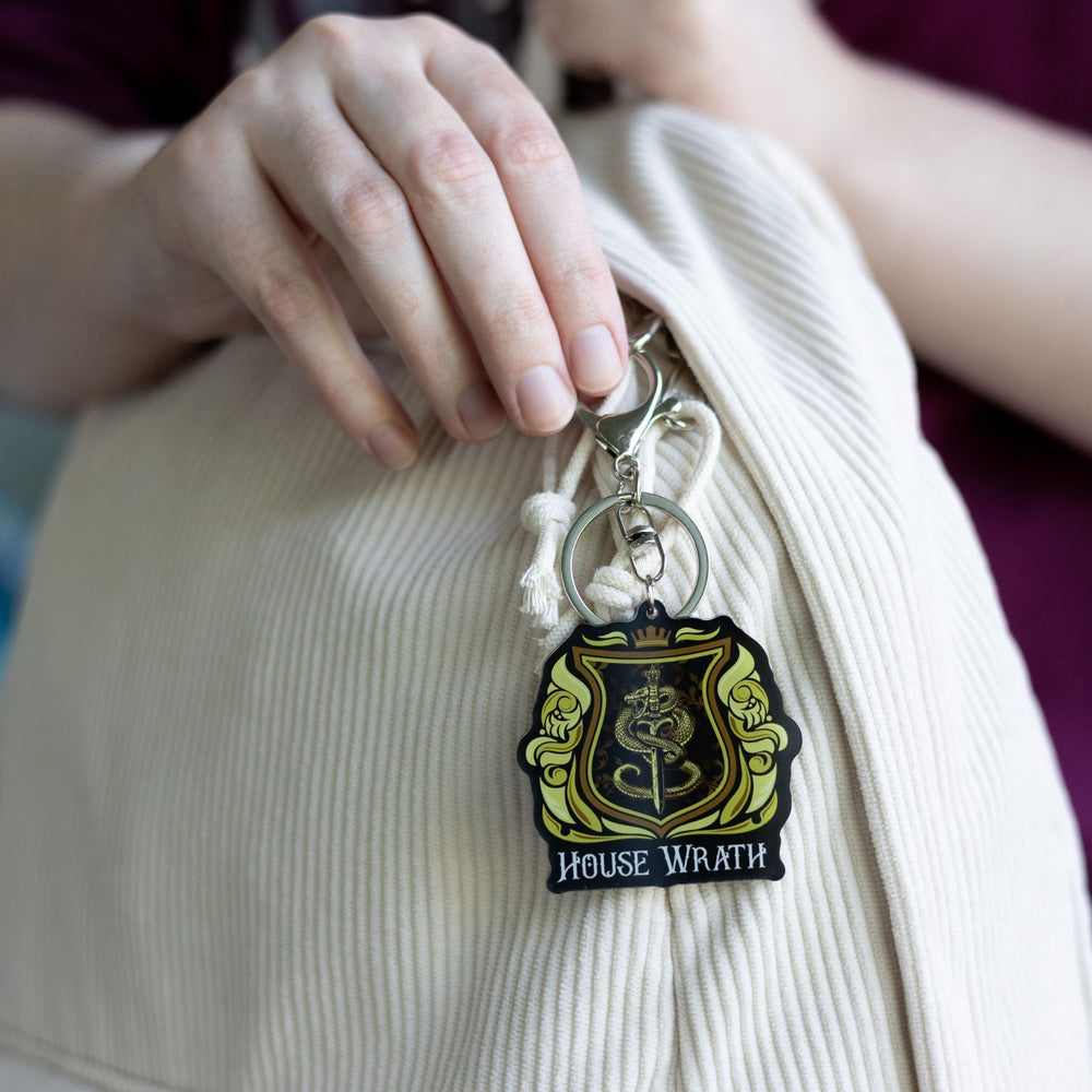 Acrylic yellow and gold keychain of a house crest with a snake around a sword and House of Wrath in white clipped to a white backpack..
