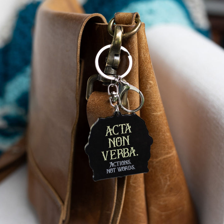  Acrylic yellow and gold keychain of a house crest that reads ‘Acta non verba. Actions not words’ clipped to a leather purse.
