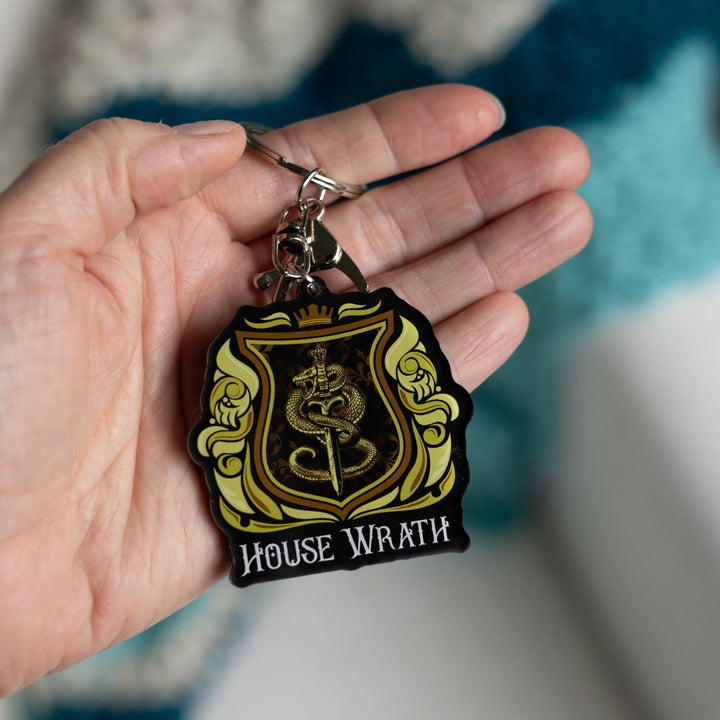  Acrylic Yellow and gold keychain of a house crest with a snake wrapped around a sword and House of Wrath in white text held in a palm.

