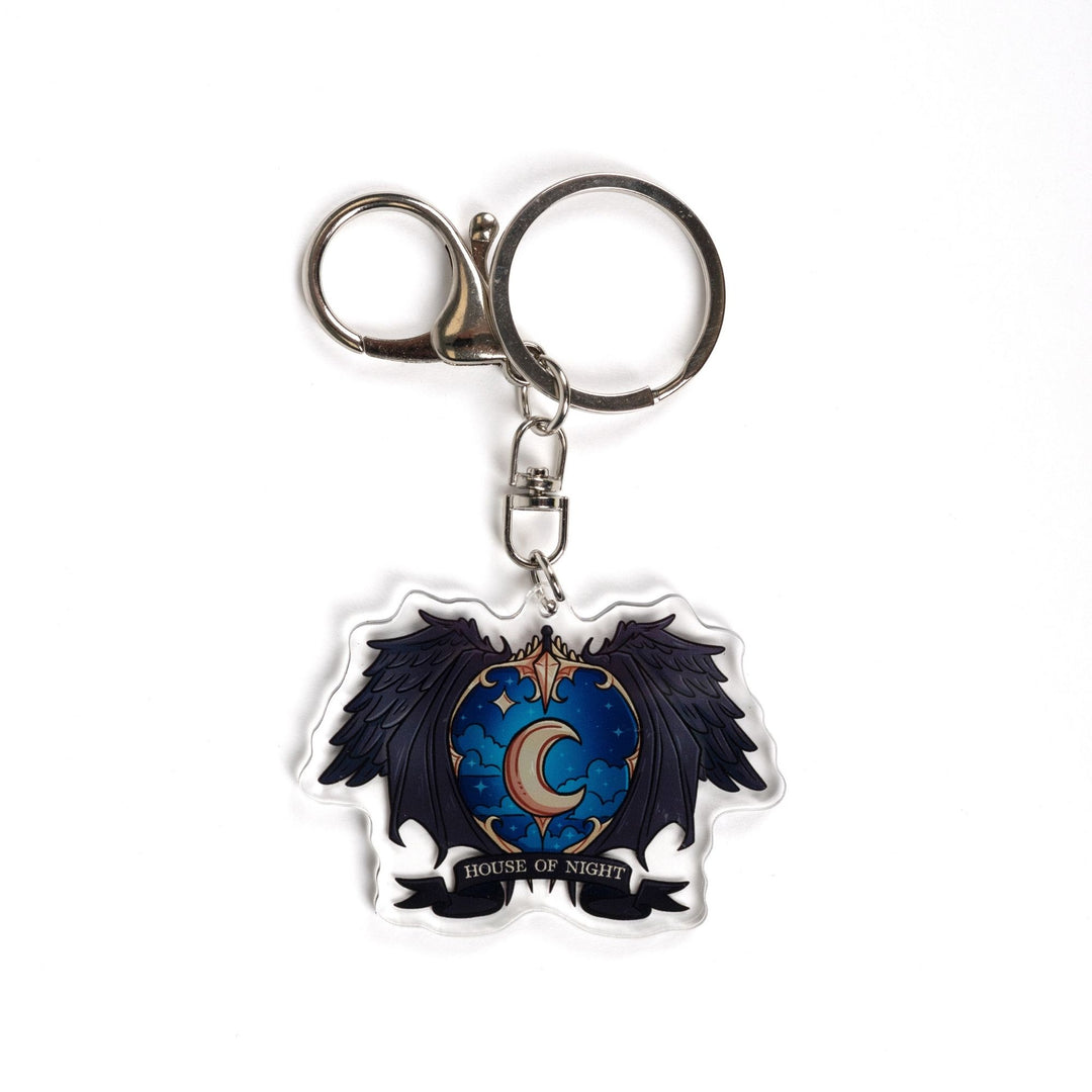 Acrylic keychain of a crest with a night sky and a gold crescent moon surrounded black wings with House of Night gold text.
