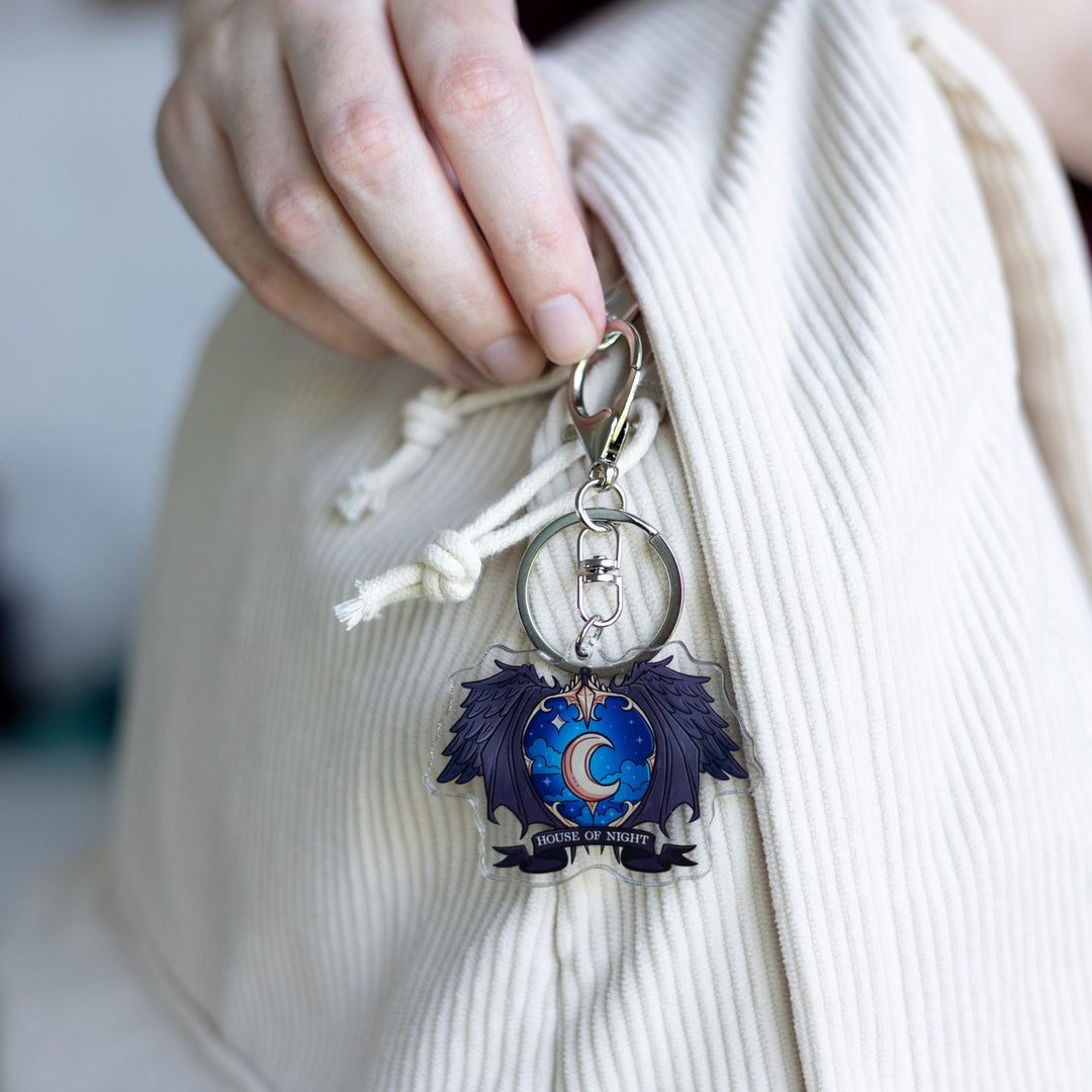 Acrylic keychain of a crest with a night sky and a gold crescent moon surrounded black wings with House of Night gold text clipped to a white backpack.
