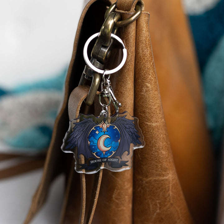  Acrylic keychain of a crest with a night sky and a gold crescent moon surrounded black wings with House of Night gold text clipped to a leather purse.
 