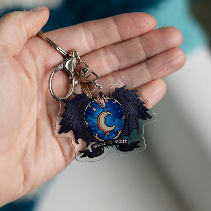 Acrylic keychain of a crest with a night sky and a gold crescent moon surrounded black wings with House of Night gold text clipped to held in a palm