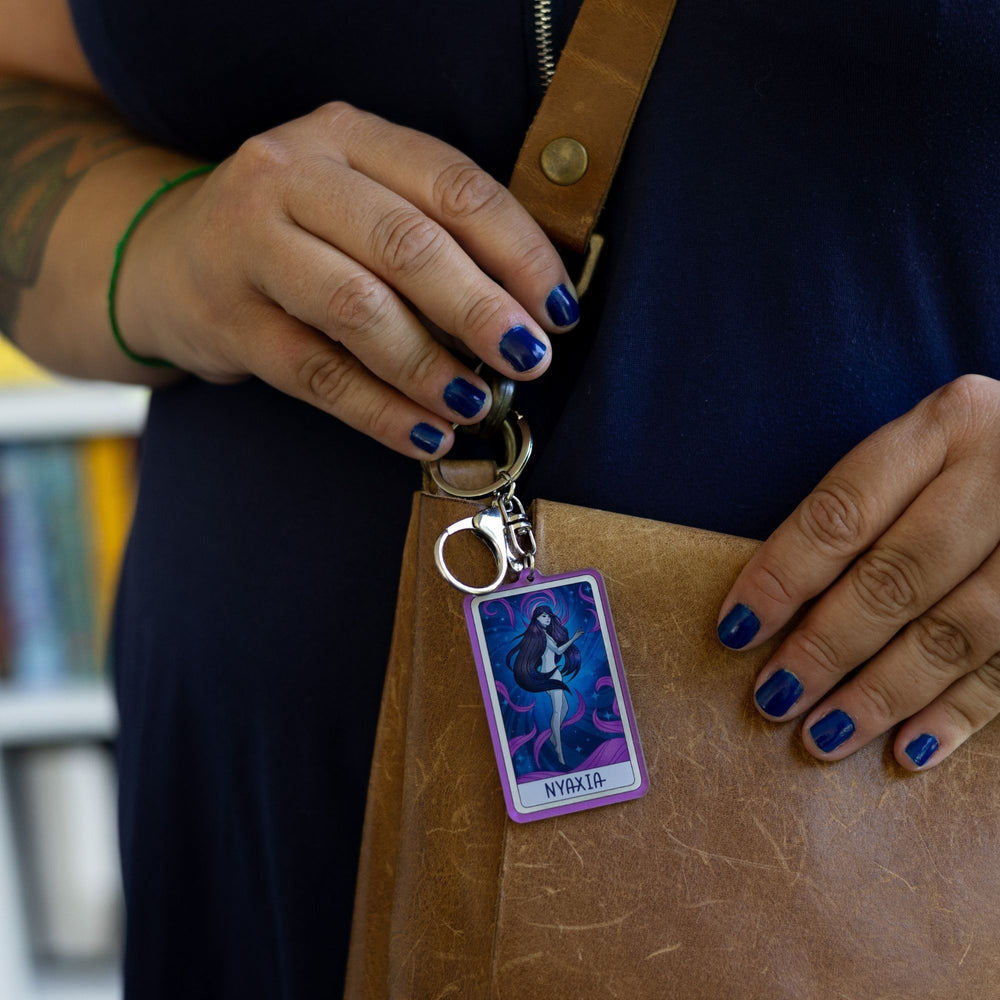 Acrylic keychain of a woman with long dark hair, black eyes floating in a night sky with purple swirls with ‘Nyaxia’ at the bottom, clipped to a leather purse. 