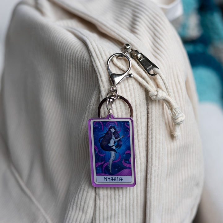 Acrylic keychain of a woman with long dark hair, black eyes floating in a night sky with purple swirls with ‘Nyaxia’ at the bottom, clipped to a white backpack.
