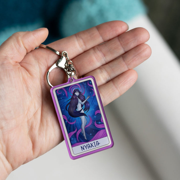 Acrylic keychain of a woman with long dark hair, black eyes floating in a night sky with purple swirls with ‘Nyaxia’ at the bottom, held in a palm.

