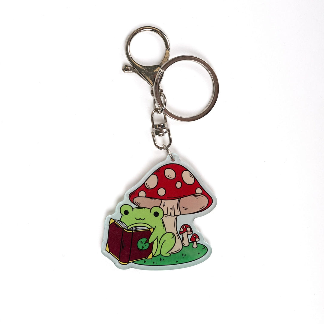 Acrylic keychain of a green frog reading a book under a mushroom.
