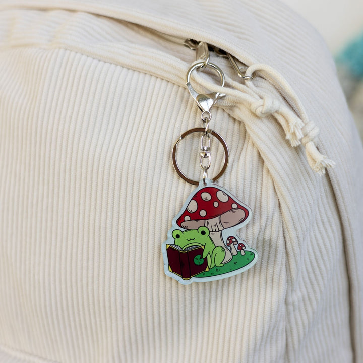  Acrylic keychain of a green frog reading a book under a mushroom clipped to a white backpack.