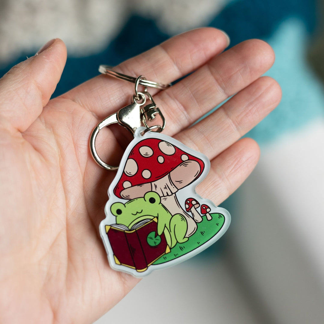 Acrylic keychain of a green frog reading a book under a mushroom held in a palm.