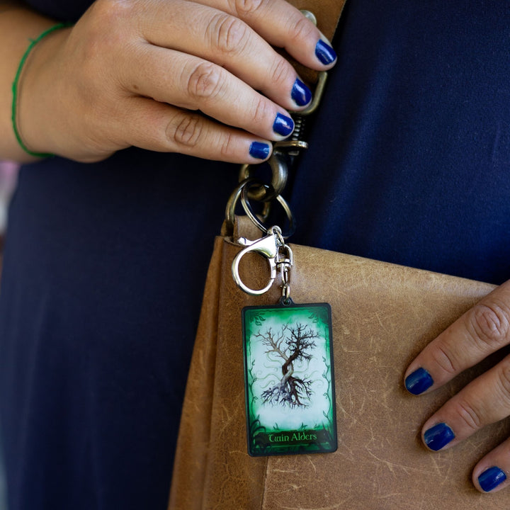 Acrylic keychain of a card with two trees intertwining on a green background. On the bottom it reads ‘Twin Alders’ clipped to a leather purse.
