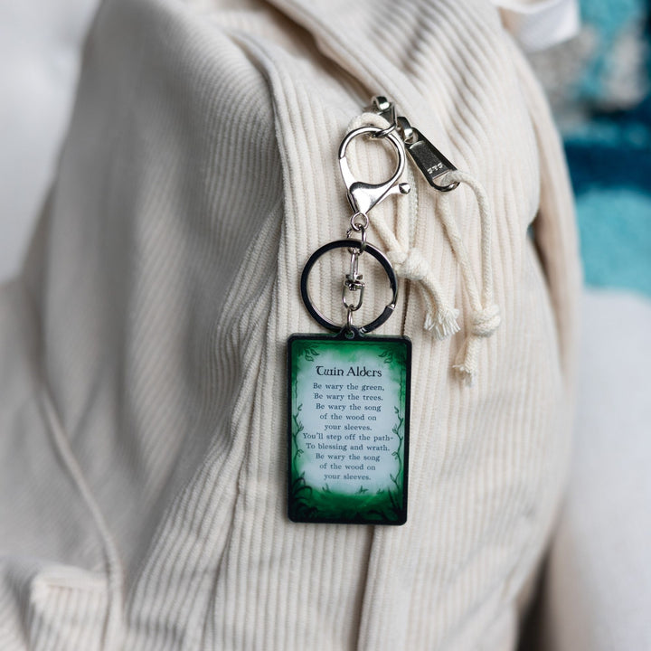 Acrylic keychain of a card that says ‘Twin Alders. Be wary the green, be wary the trees. Be wary the song of the wood on your sleeves. You’ll step off the path to blessing and wrath’  clipped to a white backpack.
