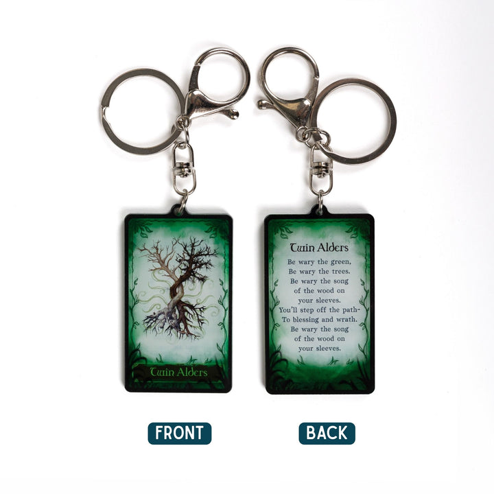 2 acrylic keychains of a tarot card. One says ‘Twin Alders’ with two trees below it. The other is a quote from One Dark Window by Rachel Gillig. 
