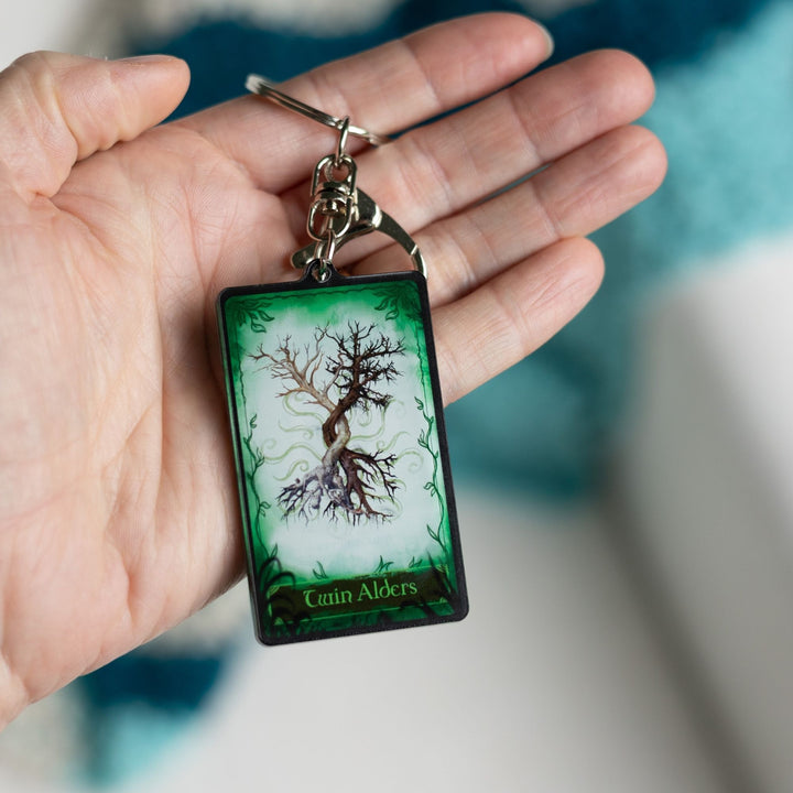 Acrylic keychain of a card with two trees intertwining on a green background. On the bottom it reads ‘Twin Alders’  held in a palm. 