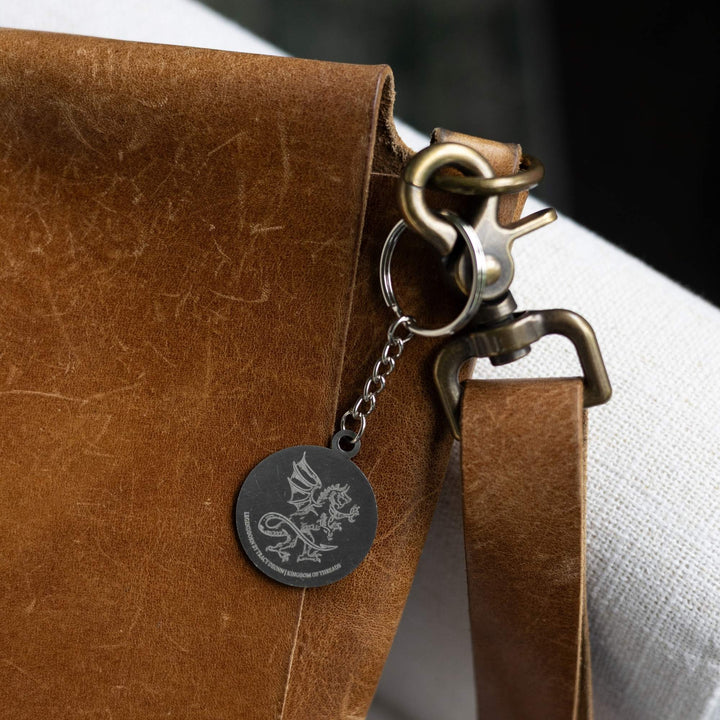 : Metal keychain of a circle with an etching of a dragon clipped to a leather purse. From Legendborn by Tracey Deonn.
