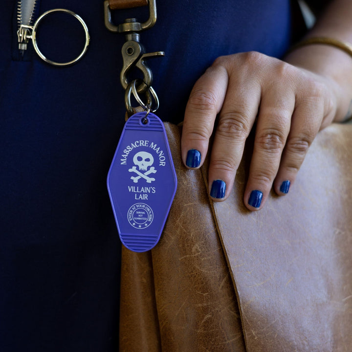 Purple plastic keychain with ‘Massacre Manor’ in white above a skull and crossbones clipped to a leather purse. From Assistant to the Villain.

