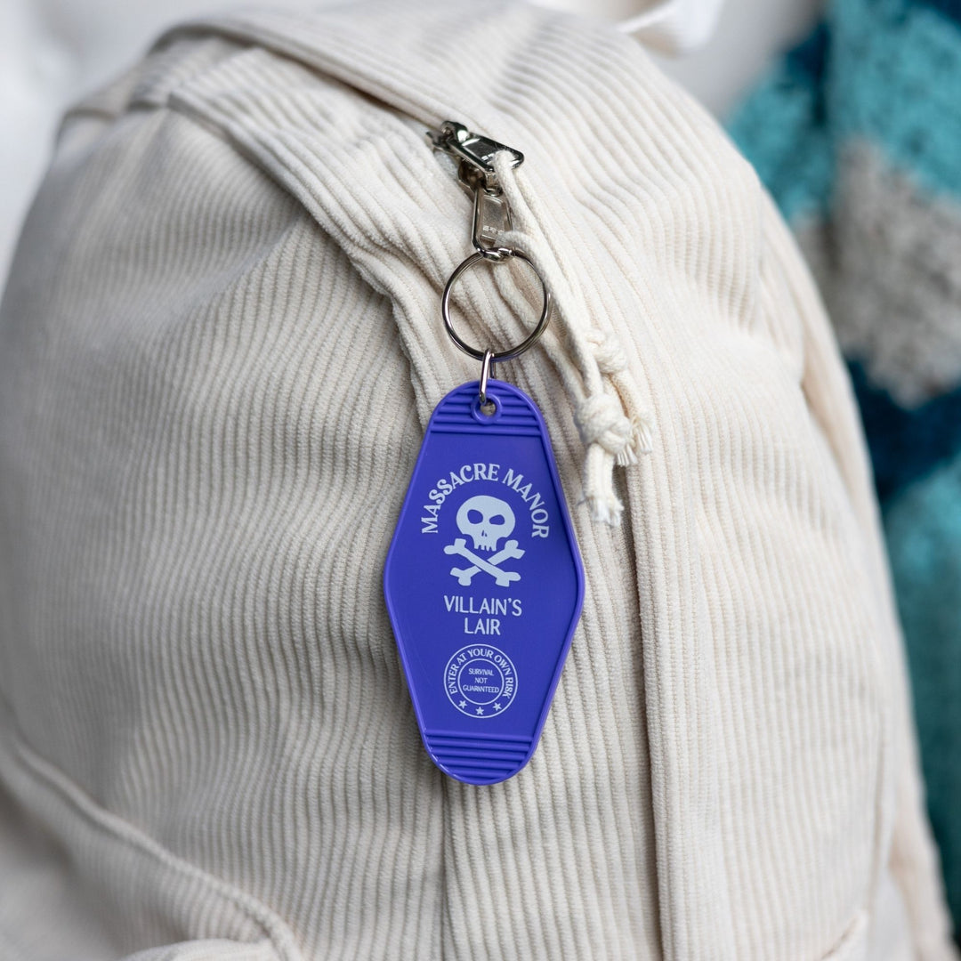 Purple plastic keychain with ‘Massacre Manor’ in white above a skull and crossbone clipped to a white backpack. From Assistant to the Villain.
