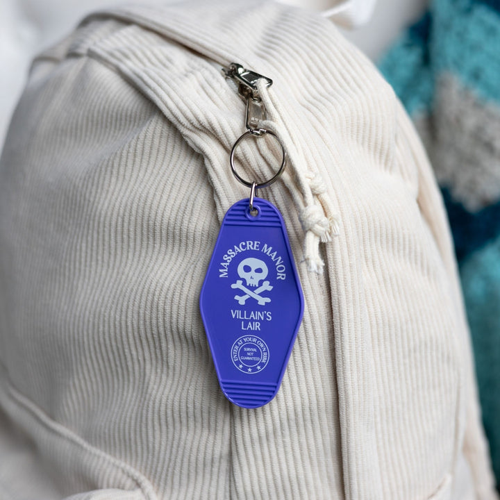 Purple plastic keychain with ‘Massacre Manor’ in white above a skull and crossbone clipped to a white backpack. From Assistant to the Villain.
