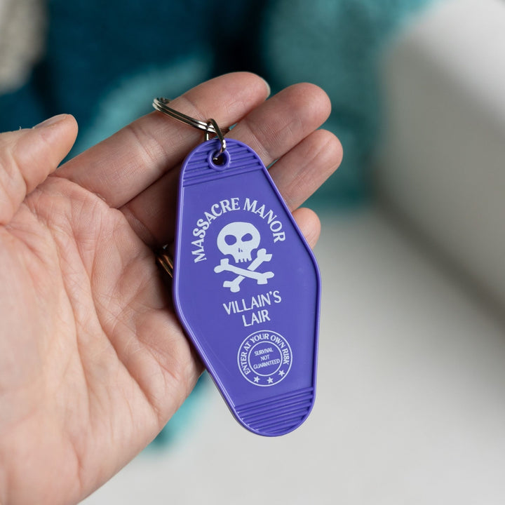  Purple plastic keychain with ‘Massacre Manor’ in white above a skull and crossbone held in a palm. From Assistant to the Villain.
