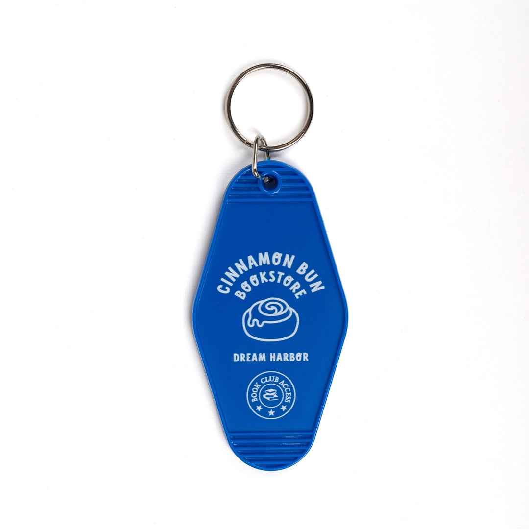 Blue plastic keychain with a white lettering and a cinnamon roll that reads ‘Cinnamon Bun Bookstore. Dream Harbor. Book club access.’ 
