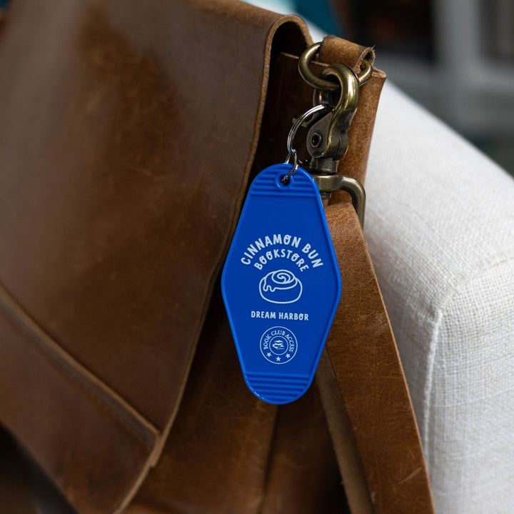 Blue plastic keychain with a white lettering and a cinnamon roll that reads ‘Cinnamon Bun Bookstore. Dream Harbor. Book club access.’ clipped to a leather purse.
