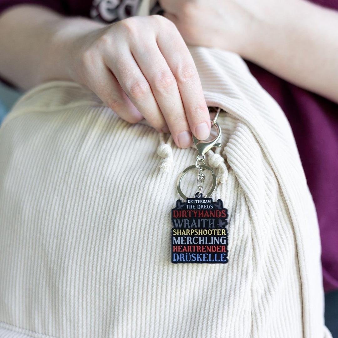 Acrylic keychain that reads ‘Ketterdam, The Dregs, Dirtyhands, Wraith, Sharpshooter, Merchling, Heartrender, Druskelle’  clipped to a white backpack.