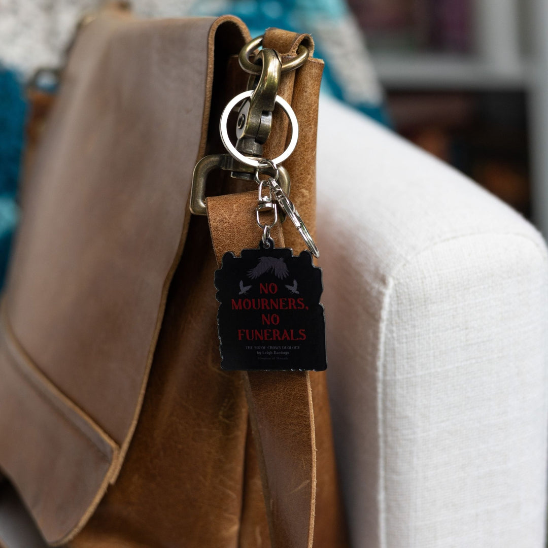 Acrylic black keychain that has ‘No mourners, no funerals’ in red text with 3 crows flying clipped to a leather purse.
