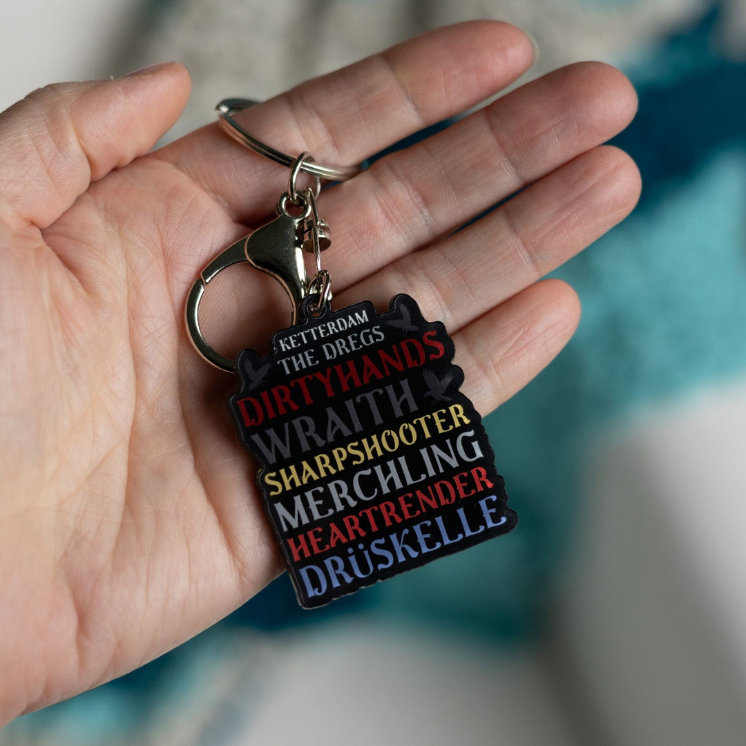 Acrylic keychain that reads ‘Ketterdam, The Dregs, Dirtyhands, Wraith, Sharpshooter, Merchling, Heartrender, Druskelle’ held in a palm.