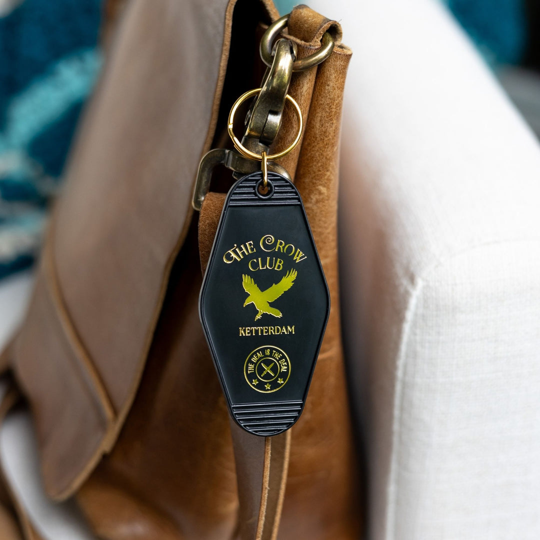 Black plastic keychain with golden lettering and a golden crow for ‘The Crow Club. Ketterdam’ clipped to a leather purse. From Six of Crows.

