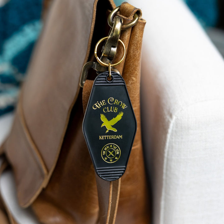 Black plastic keychain with golden lettering and a golden crow for ‘The Crow Club. Ketterdam’ clipped to a leather purse. From Six of Crows.
