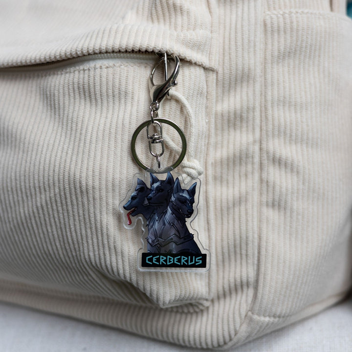 Acrylic keychain of the 3 headed dog, Cerberus in armor clipped to a white backpack. One head stoic, one head smiling, one silly with its tongue out. 