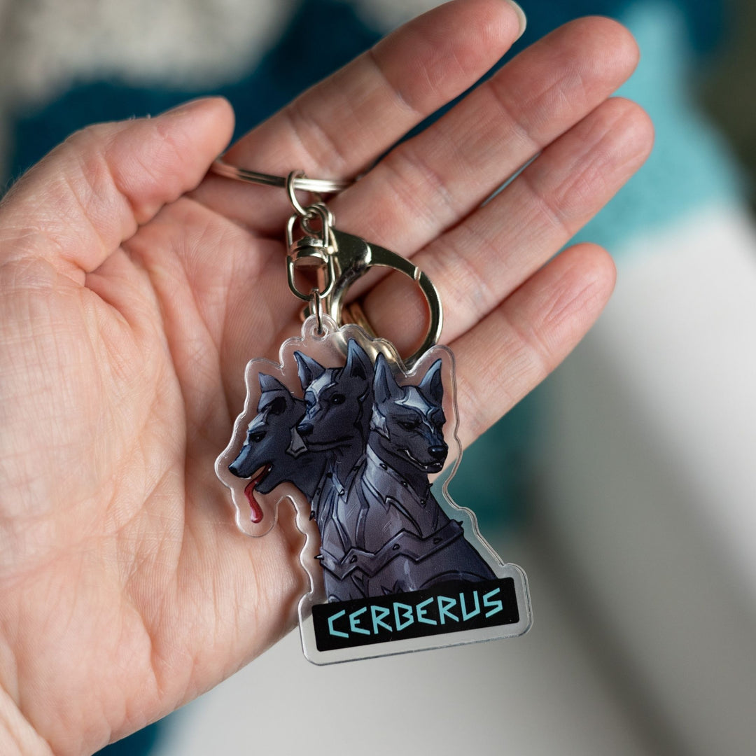 Acrylic keychain of the 3 headed dog, Cerberus in armor on a palm. One head stoic, one head smiling, one silly with its tongue out.