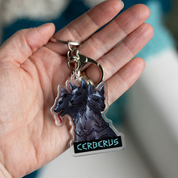 Acrylic keychain of the 3 headed dog, Cerberus in armor on a palm. One head stoic, one head smiling, one silly with its tongue out.