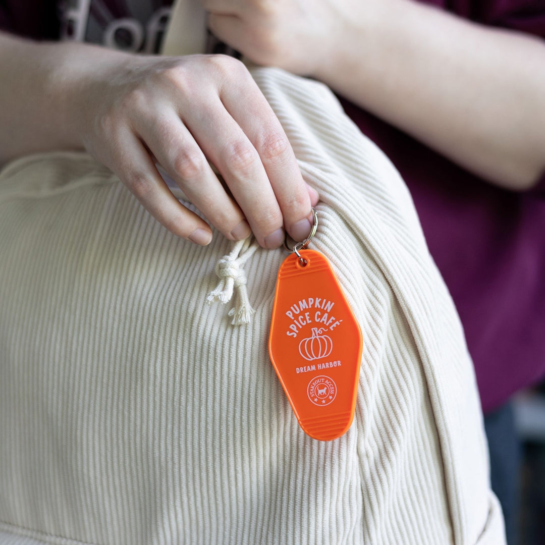 Orange motel-style keychain for ‘Pumpkin Spice Cafe. Dream Harbor’ in white letters with a pumpkin, clipped to a white backpack. From Dream Harbor.
