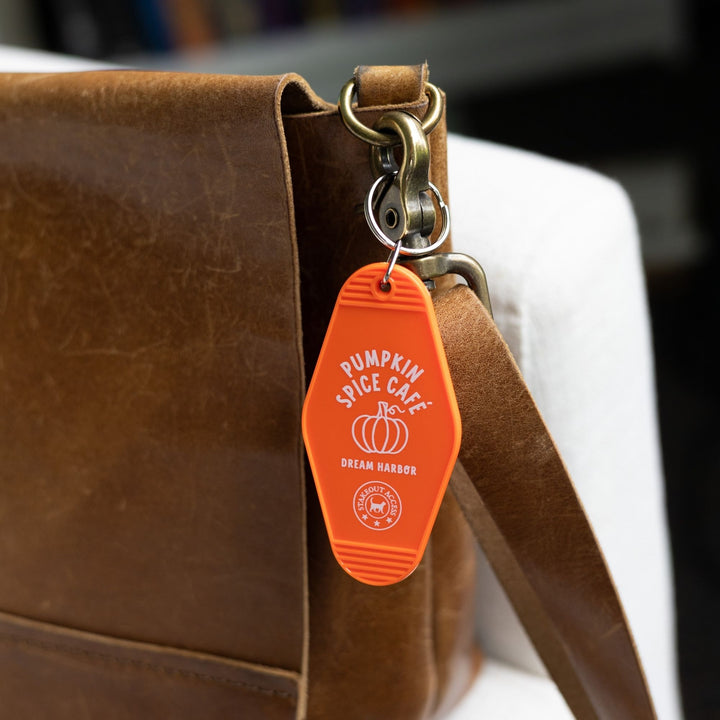 Orange motel-style keychain for ‘Pumpkin Spice Cafe. Dream Harbor’ in white letters with a pumpkin, clipped to a leather purse. From Dream Harbor.