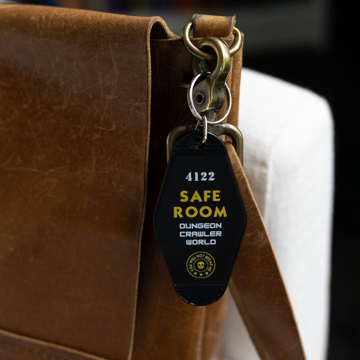 Black motel-style keychain with white and yellow lettering that reads ‘4112 Safe Room Dungeon Crawler World’ clipped to a leather purse. 
