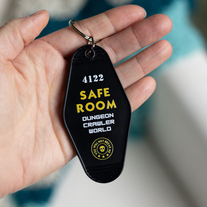 Black motel-style keychain with white and yellow lettering that reads ‘4112 Safe Room Dungeon Crawler World’held in a palm. From Dungeon Crawler Carl.
