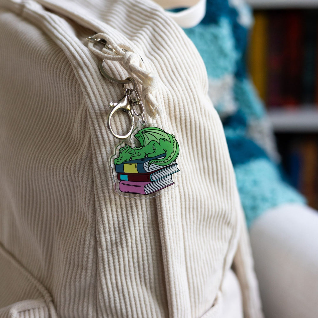 Acrylic keychain of a small green dragon asleep on a pile of books clipped to a white backpack.