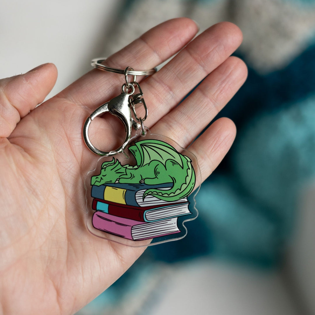  Acrylic keychain of a small green dragon asleep on a pile of books held in a palm. 
