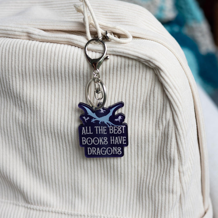 Purple acrylic keychain of one big and three small flying dragons that reads ‘All the best books have dragons’ in white letters, clipped to a white backpack.

