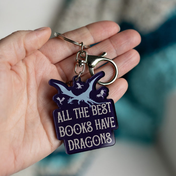 Purple acrylic keychain of one big and three small flying dragons that reads ‘All the best books have dragons’ in white letters, held in a palm.