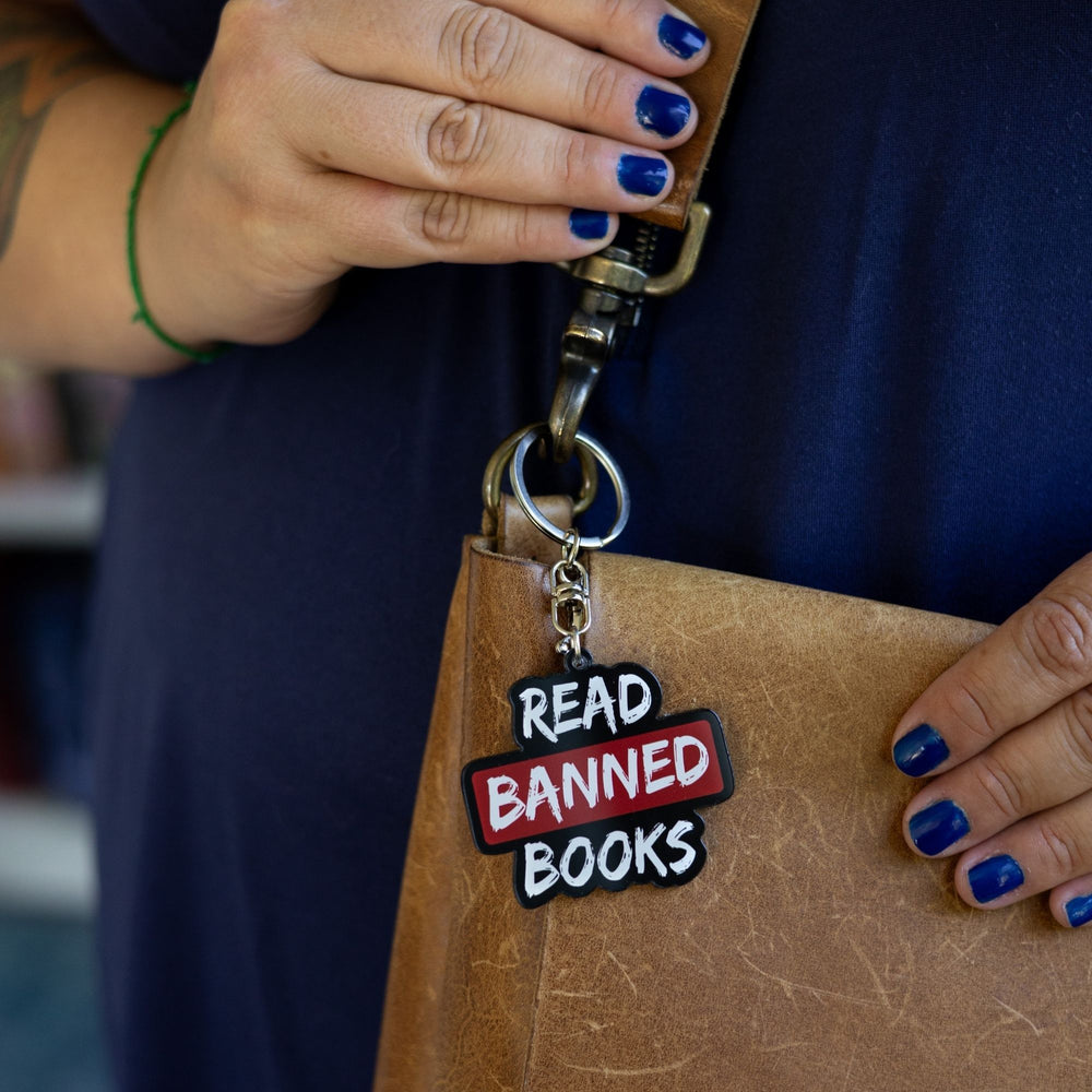 Acrylic keychain that says ‘Read banned books’ in white lettering outlined in black. The word banned is red clipped to a leather purse.