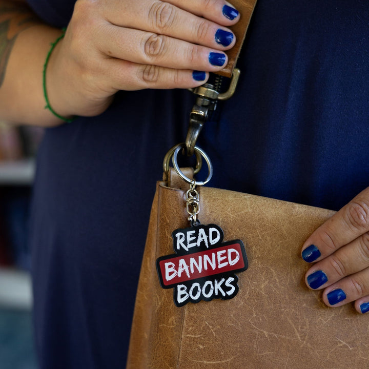 Acrylic keychain that says ‘Read banned books’ in white lettering outlined in black. The word banned is red clipped to a leather purse.