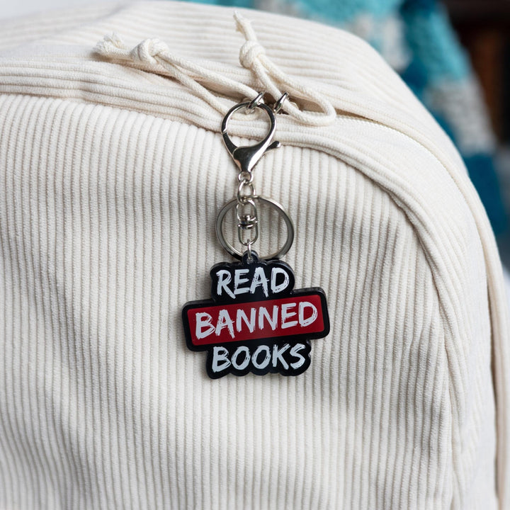  Acrylic keychain that says ‘Read banned books’ in white lettering outlined in black. The word banned is red clipped to a white backpack.