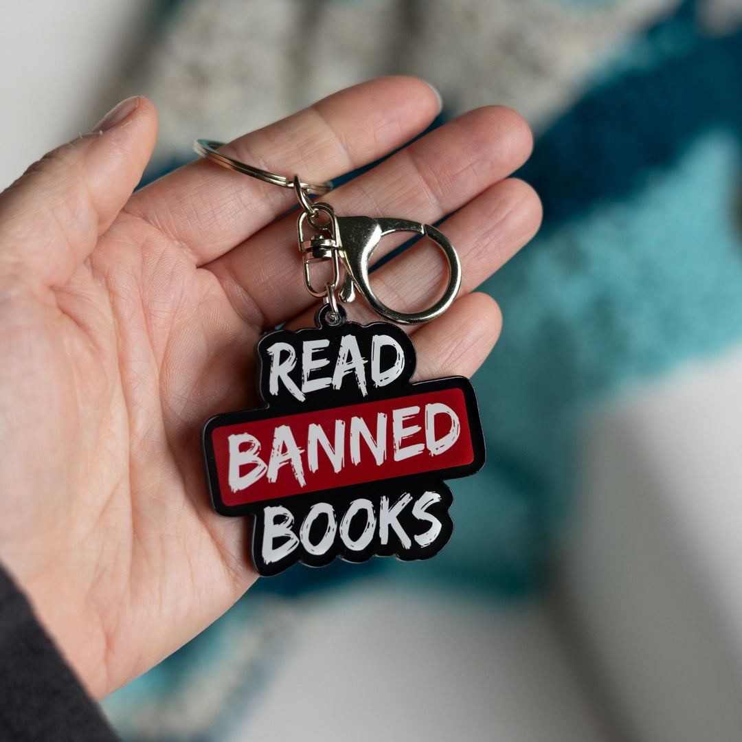 Acrylic keychain that says ‘Read banned books’ in white lettering outlined in black. The word banned is red held in a palm.